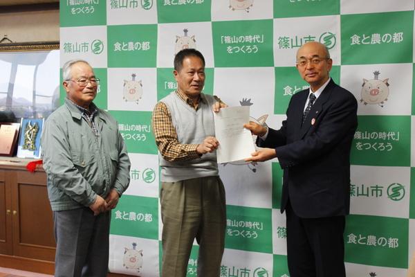 市長が田中 哲夫先生から、市民や行政が取り組む具体策の案を手渡しでもらっており、メガネをかけた白髪の男性も一緒に写っている写真